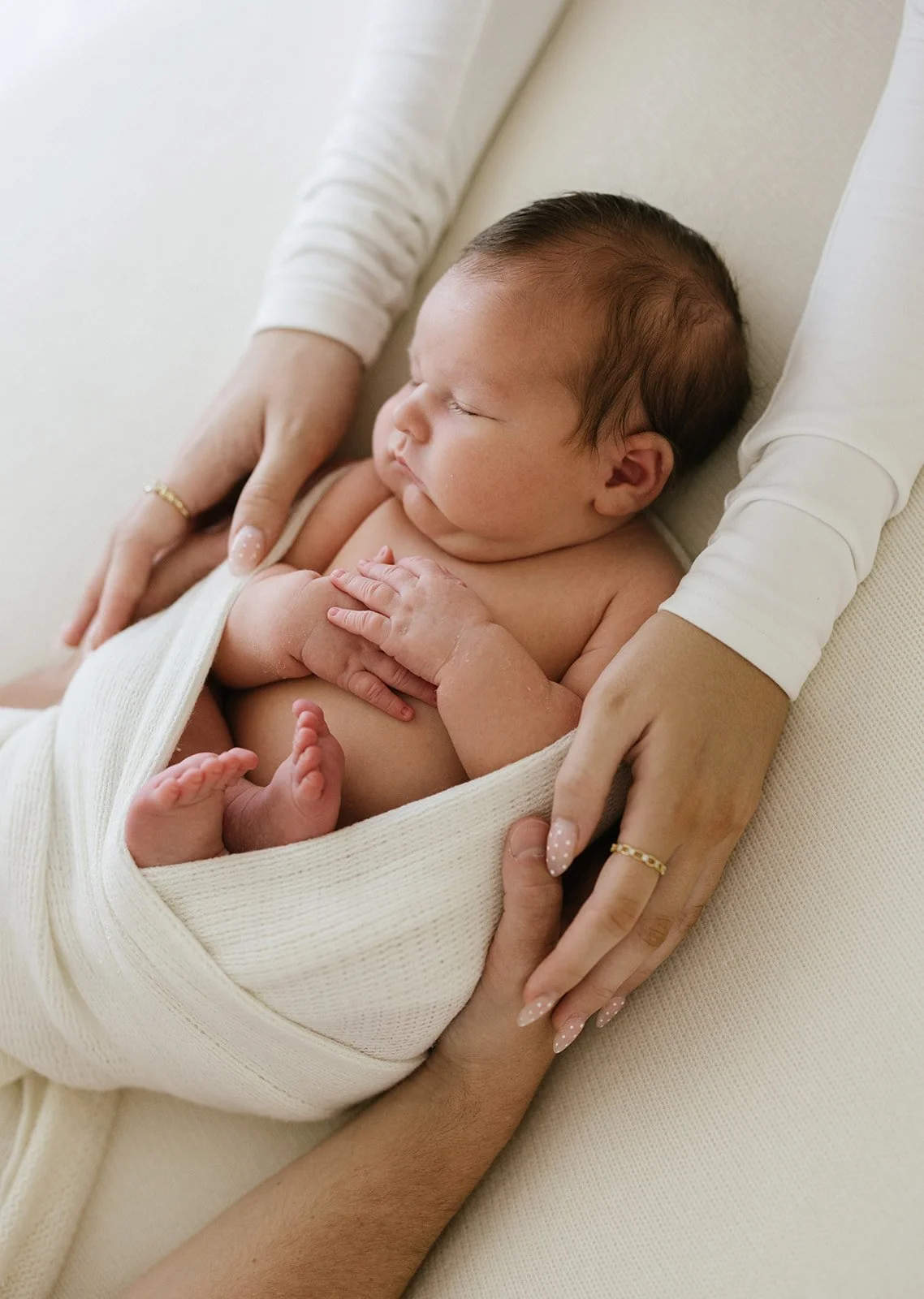 A sleeping newborn baby held gently by an adult, wrapped in a cream-colored blanket, with hands and arms visible supporting the baby.