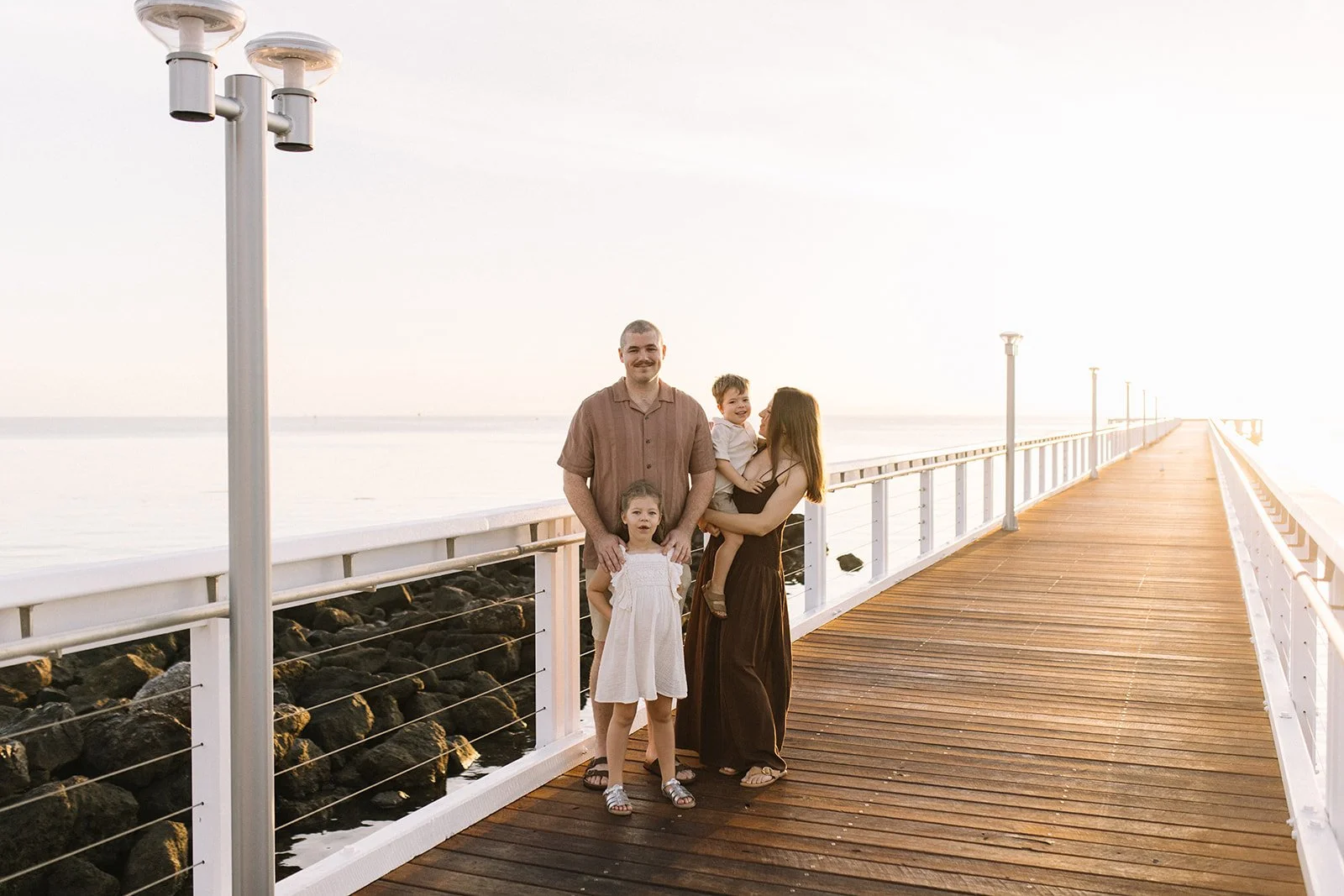 A family of four stands on a wooden pier over water during sunset, with rocks below the pier and lampposts lining the walkway. Taken by Wylde Folk Studio, Brisbane. Family photographer.