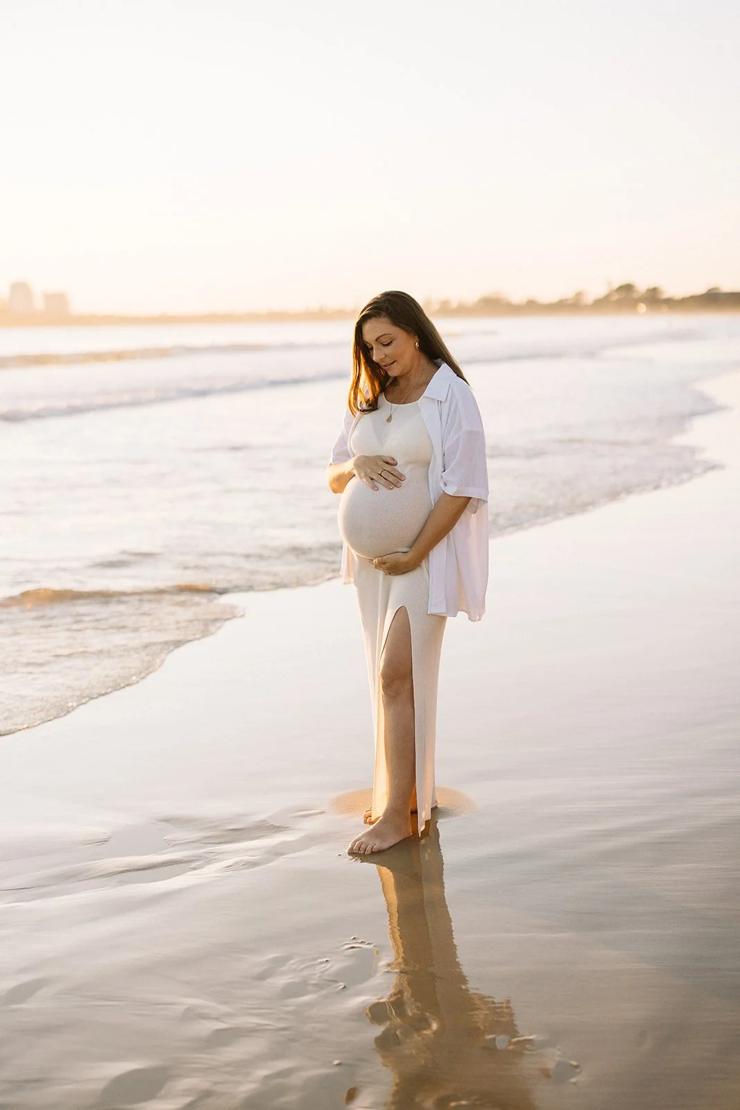 Pregnant woman in a white dress and shirt standing on the beach at sunset, with her hands on her belly and smiling. Taken by Wylde Folk Studio, Brisbane. Maternity photographer.