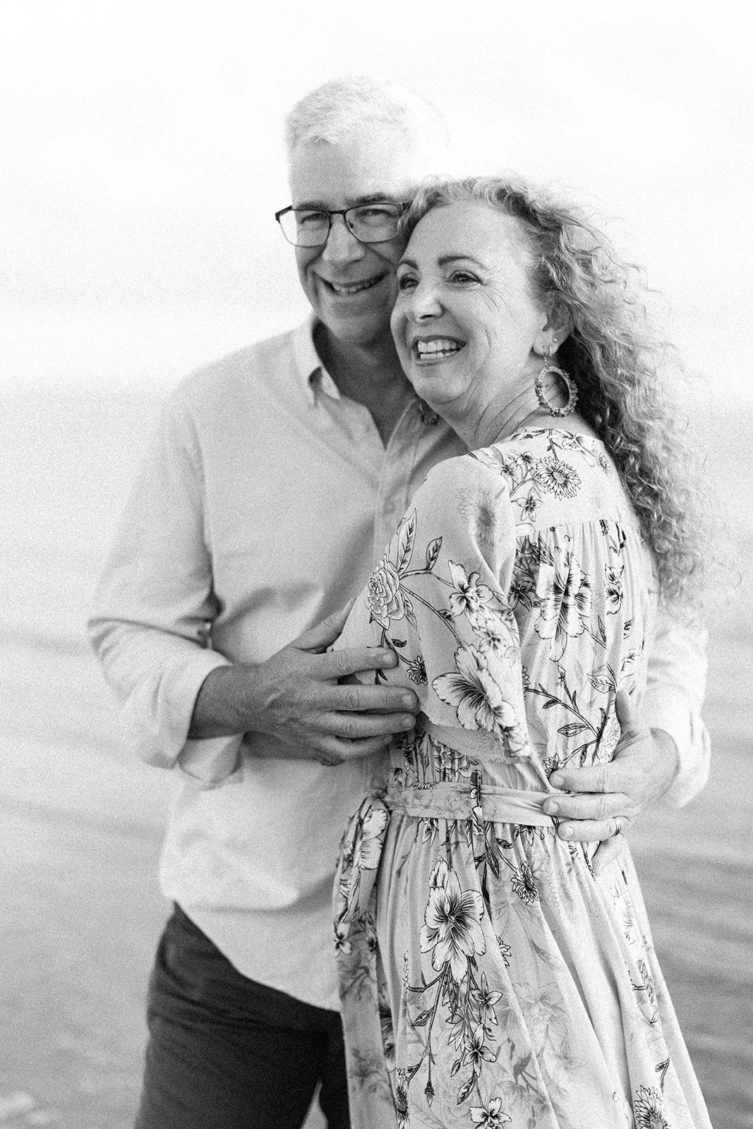 A smiling older man and woman embracing each other outdoors, woman in a floral dress and large hoop earrings, man wearing glasses and a light shirt. Taken by Wylde Folk Studio, Brisbane. Family photographer.