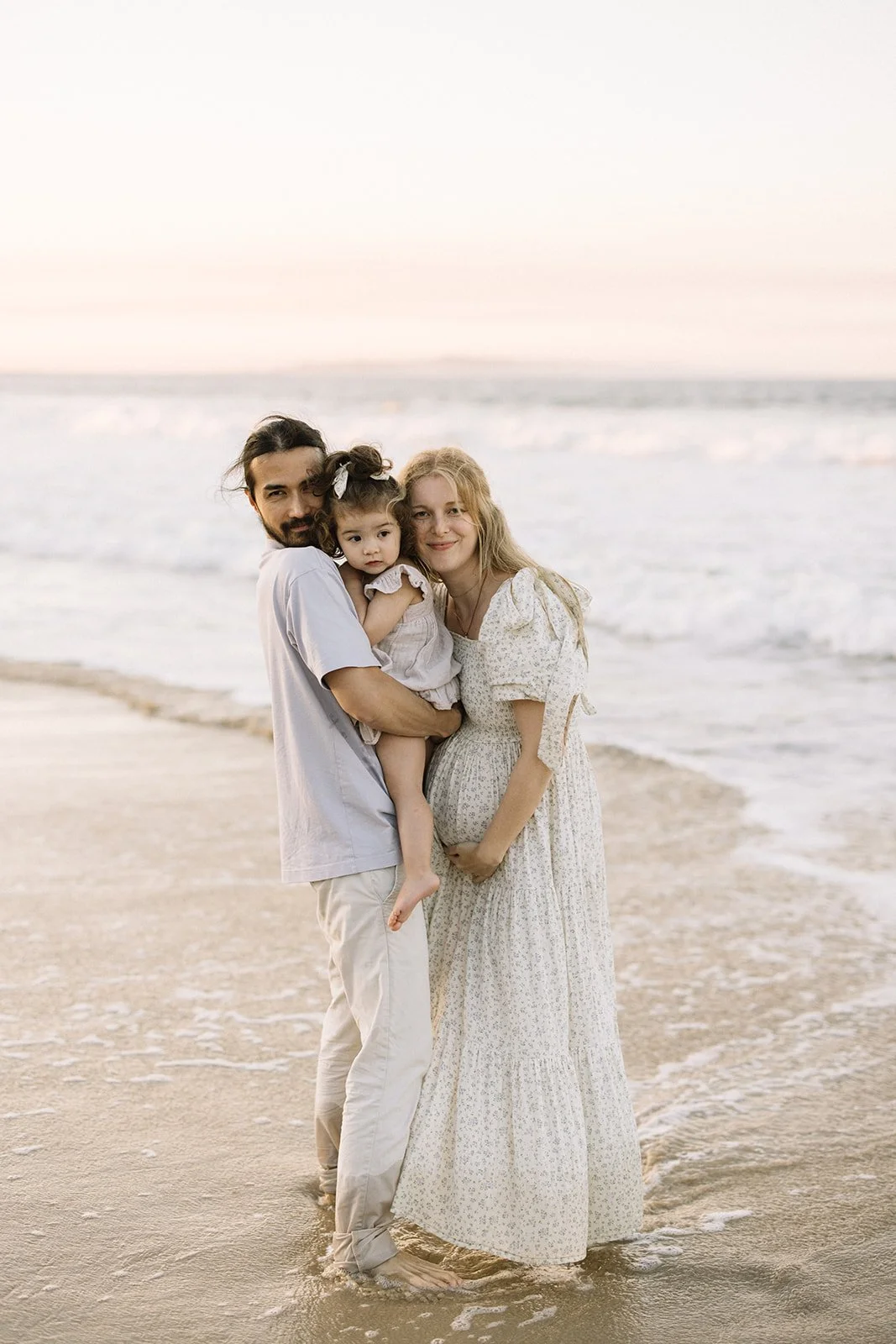 A family of three standing on the beach at sunset, with the ocean in the background. The man is holding a young girl, and a woman, possibly pregnant, is standing beside them.