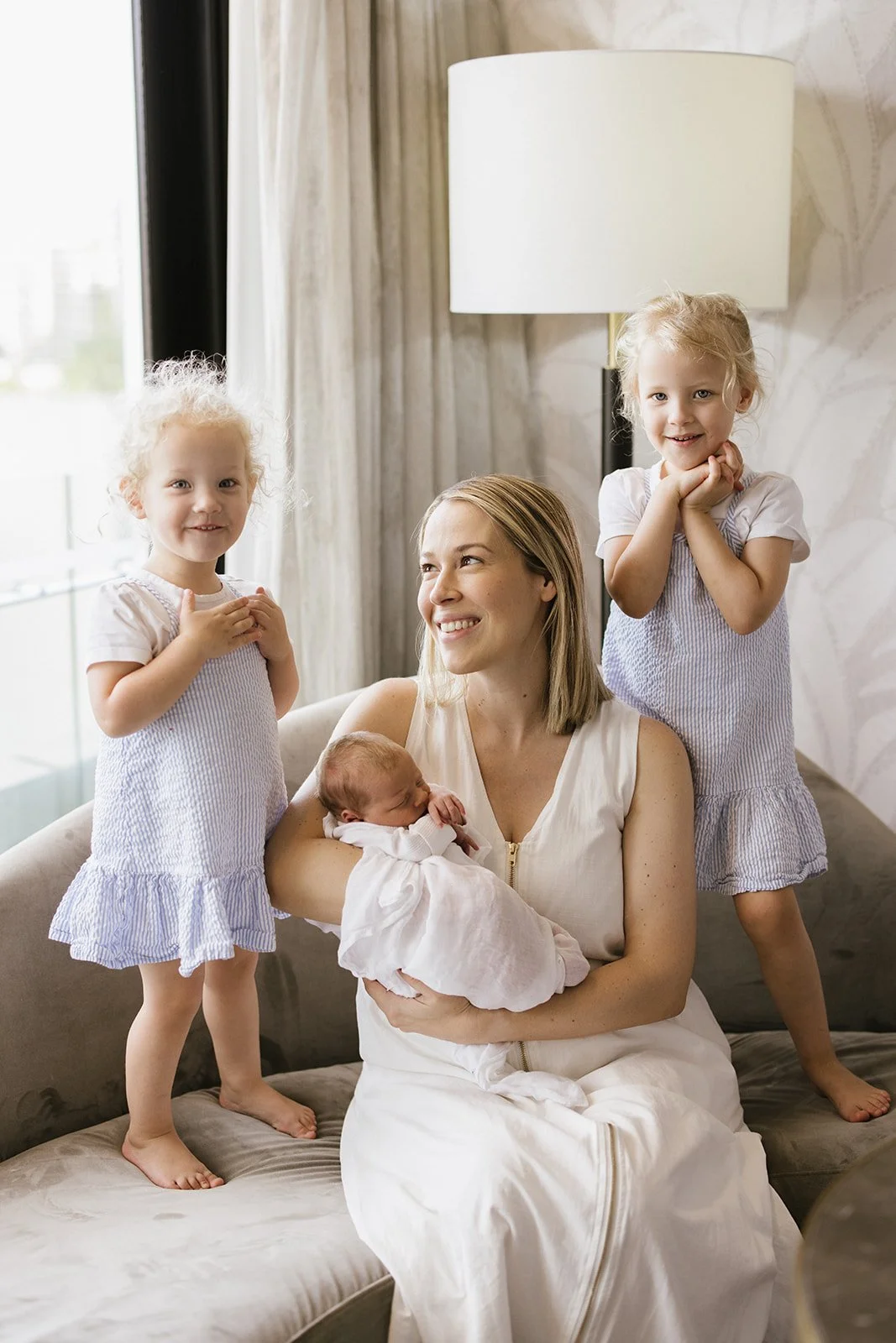 A woman with three young girls, one holding a newborn baby, all smiling and sitting on a sofa in a bright living room.