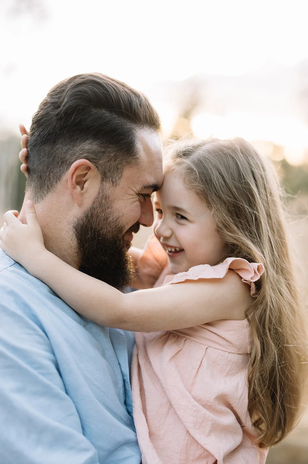 A man hugging a young girl outside during sunset, both smiling and touching foreheads. Taken by Wylde Folk Studio, Brisbane. Family photographer.