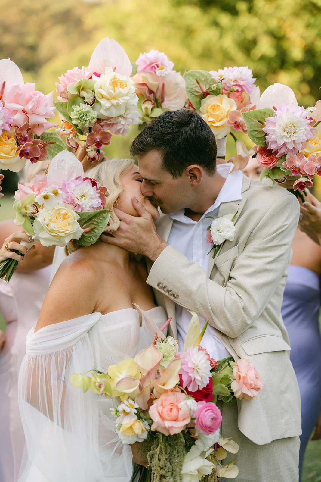 A bride and groom are kissing during their wedding, surrounded by floral bouquets and an arch of colorful flowers outdoors.