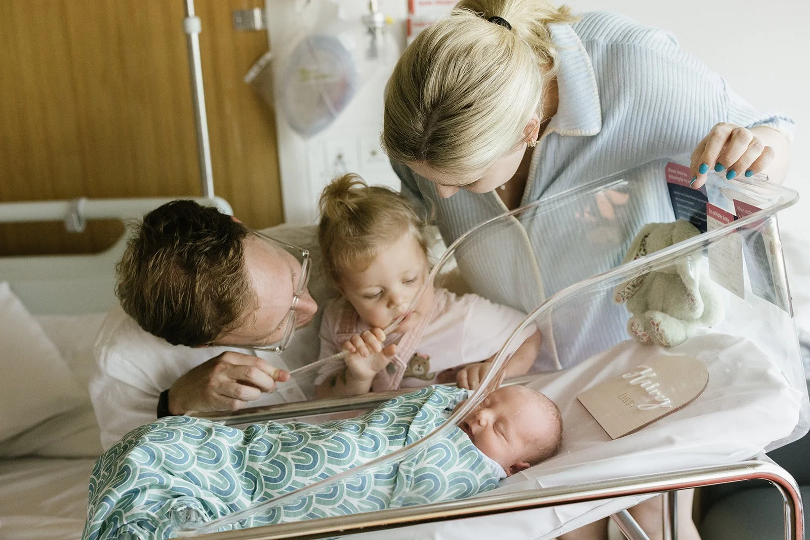 A family at a hospital bedside, with a mother, father, and two young children, as they meet their newborn baby in an incubator.