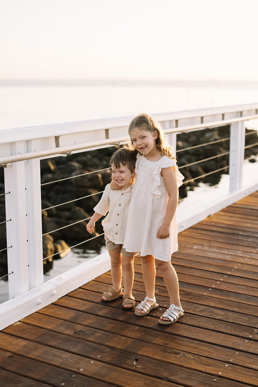Two young children, a boy and a girl, standing on a wooden dock by the water, smiling and enjoying each other's company during sunset. Taken by Wylde Folk Studio, Brisbane. Family photography.