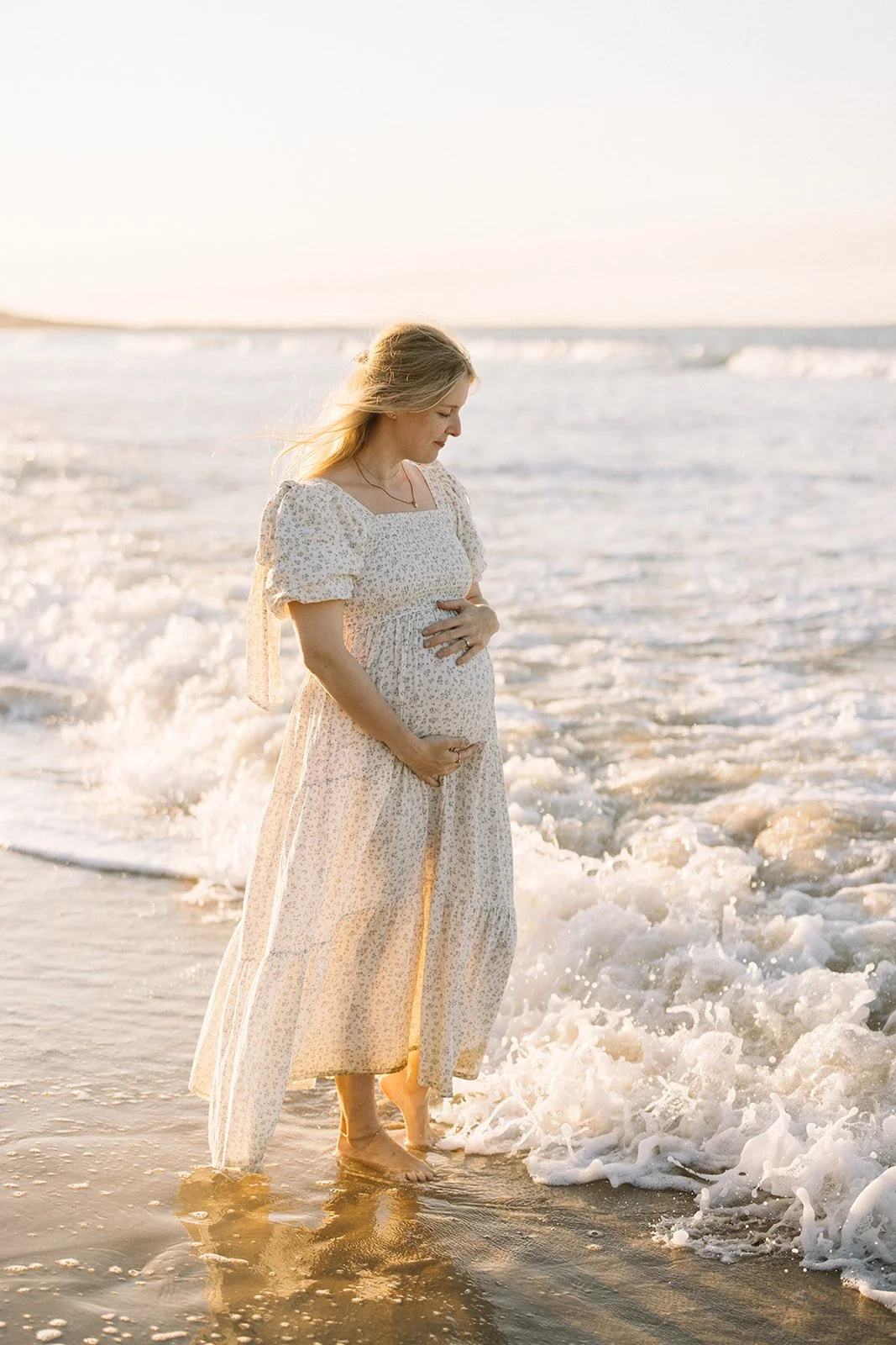 Pregnant woman in a white floral dress standing in shallow ocean waves at sunset, touching her belly, with a serene expression. Taken by Wylde Folk Studio, Brisbane. Maternity photography.