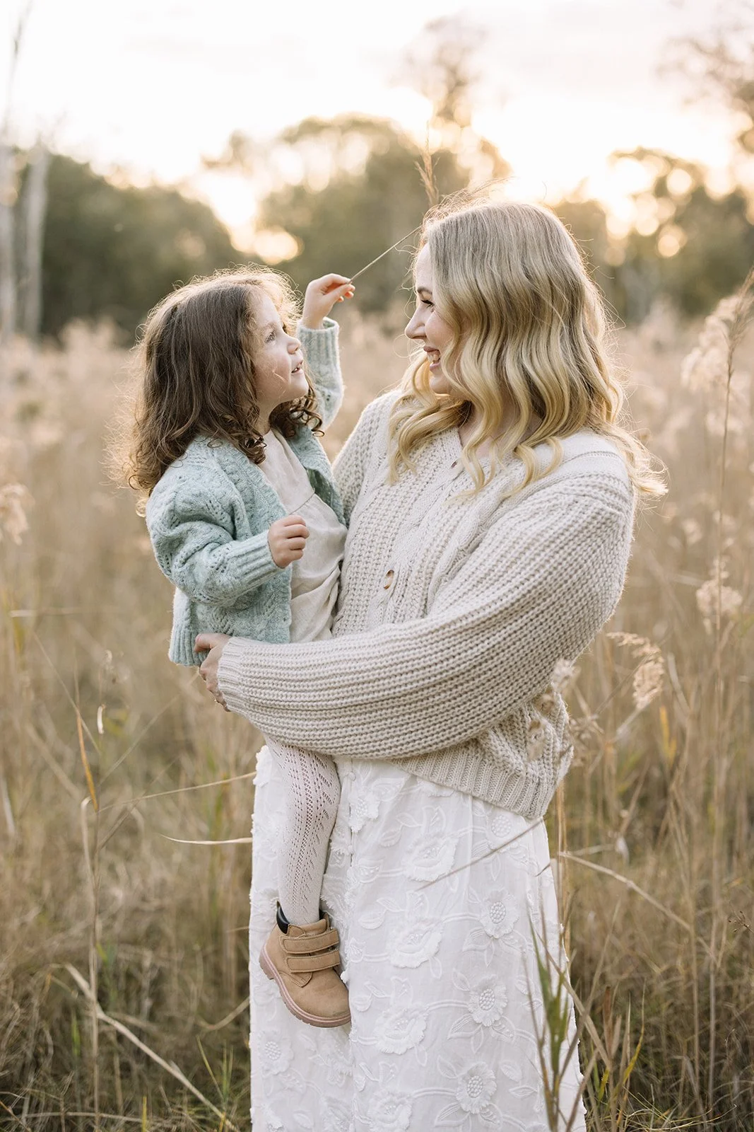A woman holding a young girl in a field of tall grass during sunset, both smiling at each other. The girl is waving a dandelion, and the woman is wearing a beige knitted sweater, while the girl is dressed in a light cardigan and dress.