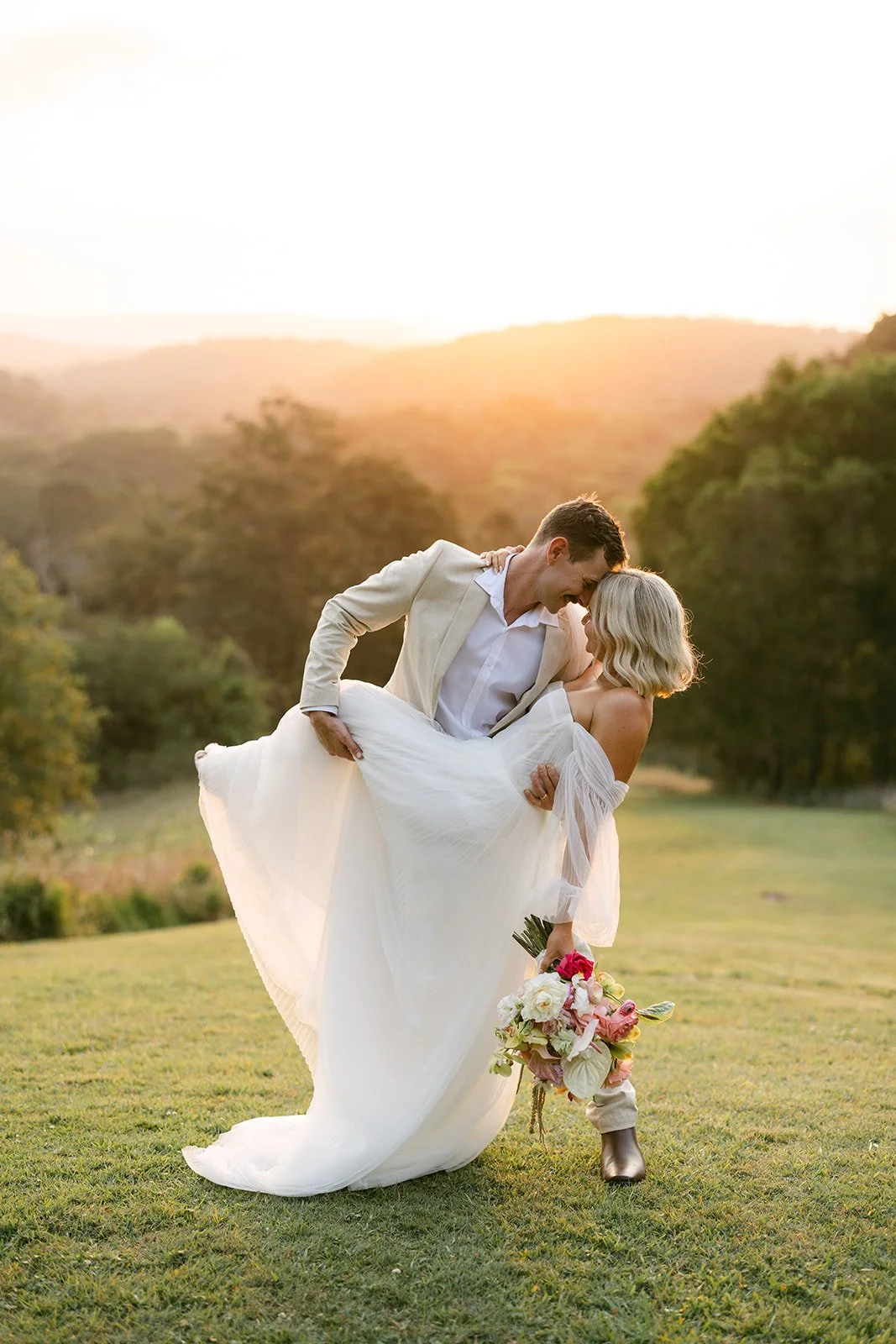 A bride and groom sharing a dance outdoors at sunset, with the groom lifting the bride and her holding a bouquet of flowers.
