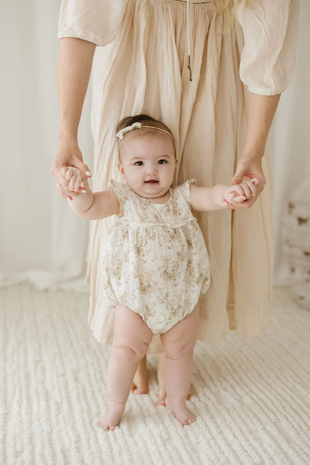 A baby girl in a floral dress is holding hands with an adult, standing on a beige carpet in a bright room. Taken at Wylde Folk Studio - Brisbane baby sitting photography.