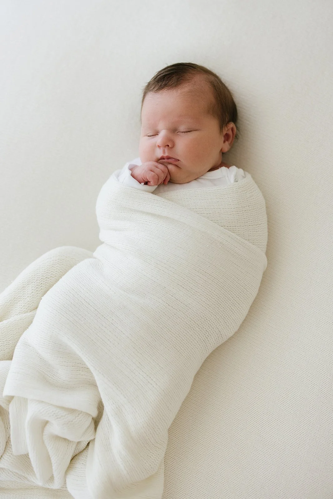 A sleeping newborn baby swaddled in a white blanket, lying on a white surface.
