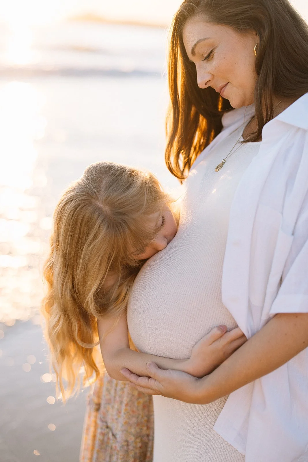 A pregnant woman with long, dark hair stands near a body of water during sunset, holding her belly. A young girl with long, red hair is hugging her belly and resting her head on it.