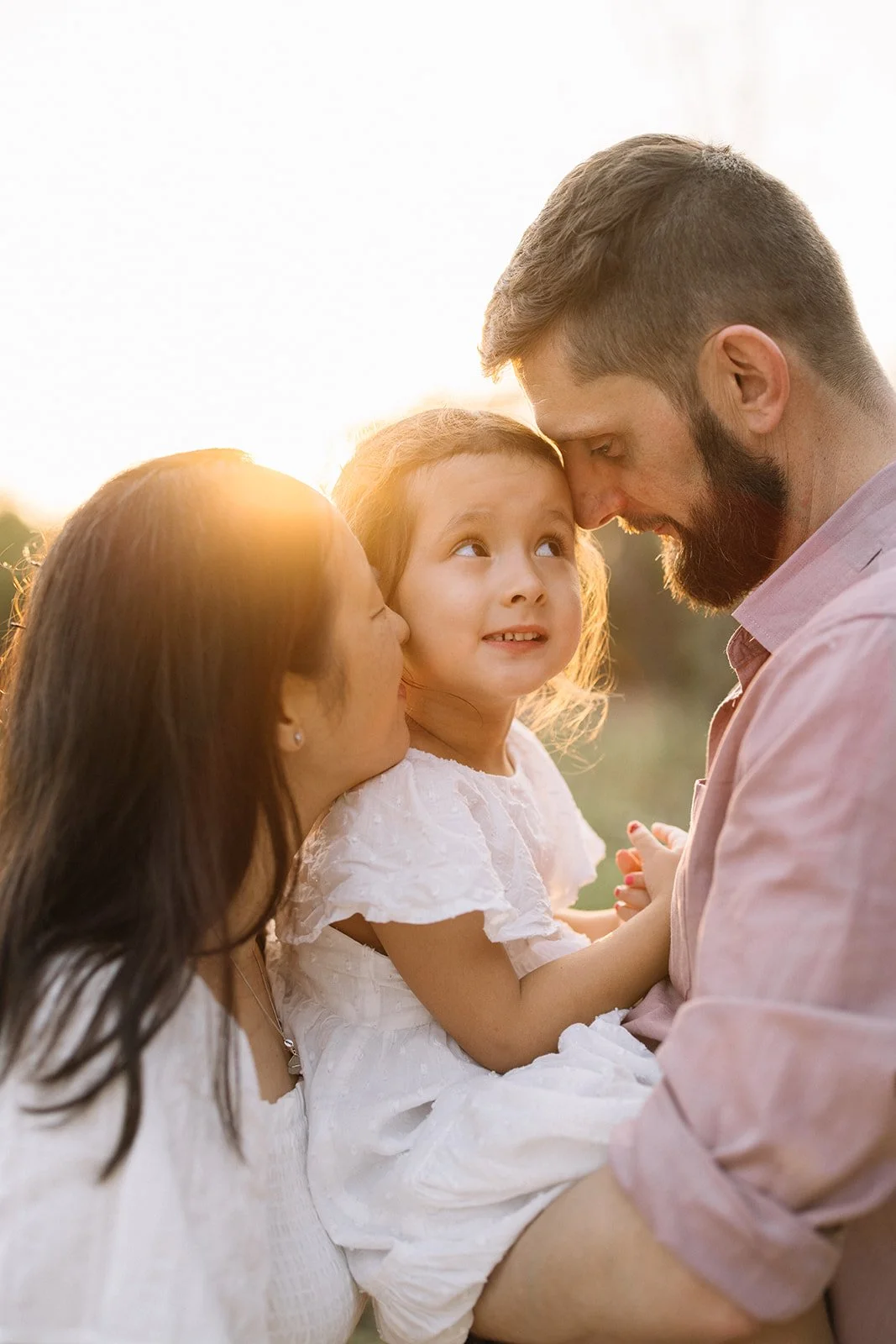 A family of three, including a little girl, a woman, and a man, outdoors during sunset, with the parents holding the girl and all touching foreheads. Taken by Wylde Folk Studio, Brisbane. Family photographer.