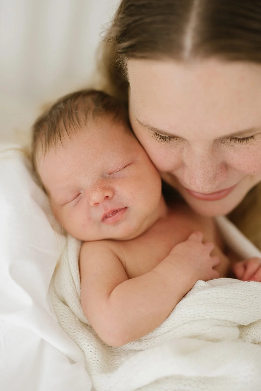 A newborn baby with light brown hair sleeps with mother cuddling into them