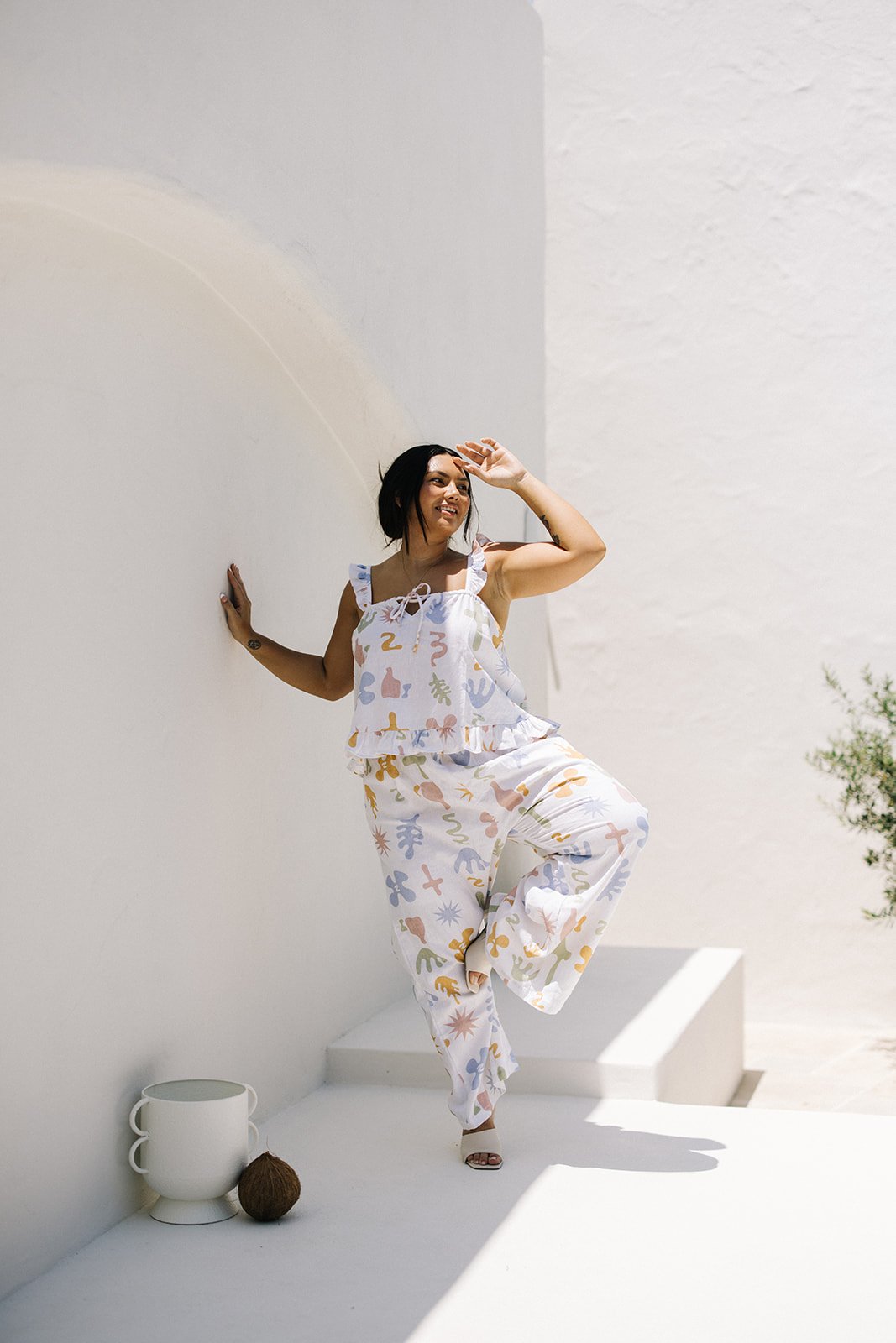 a woman with dark hair leans against a white wall posing for a commercial branding shoot for a woman's clothing label