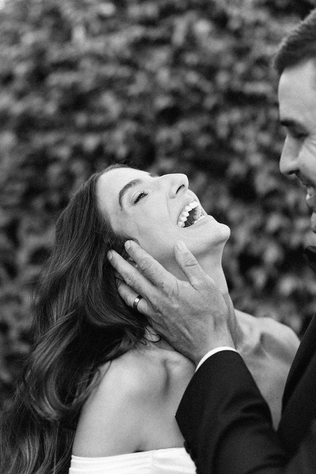 A woman with long hair smiling and laughing, being touched gently on the face by a man in a suit, outdoors with blurred background.