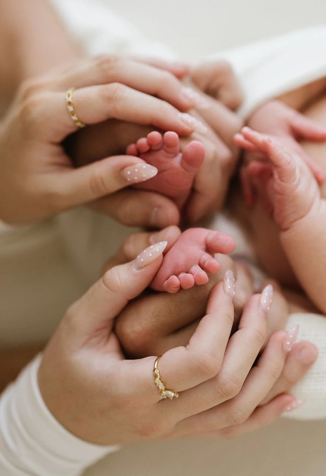 Close-up of a newborn baby's small feet being gently held by adults' hands, with the adults' nails painted with white polish and wearing gold rings.