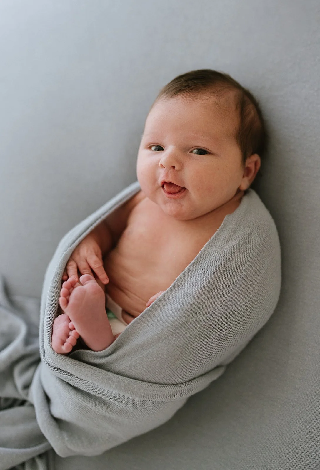 Close-up of a newborn baby wrapped in a gray blanket, lying on a soft gray surface. The baby is looking at the camera with a slight smile, one hand near the chest and the other near the feet. Brisbane newborn photographer.