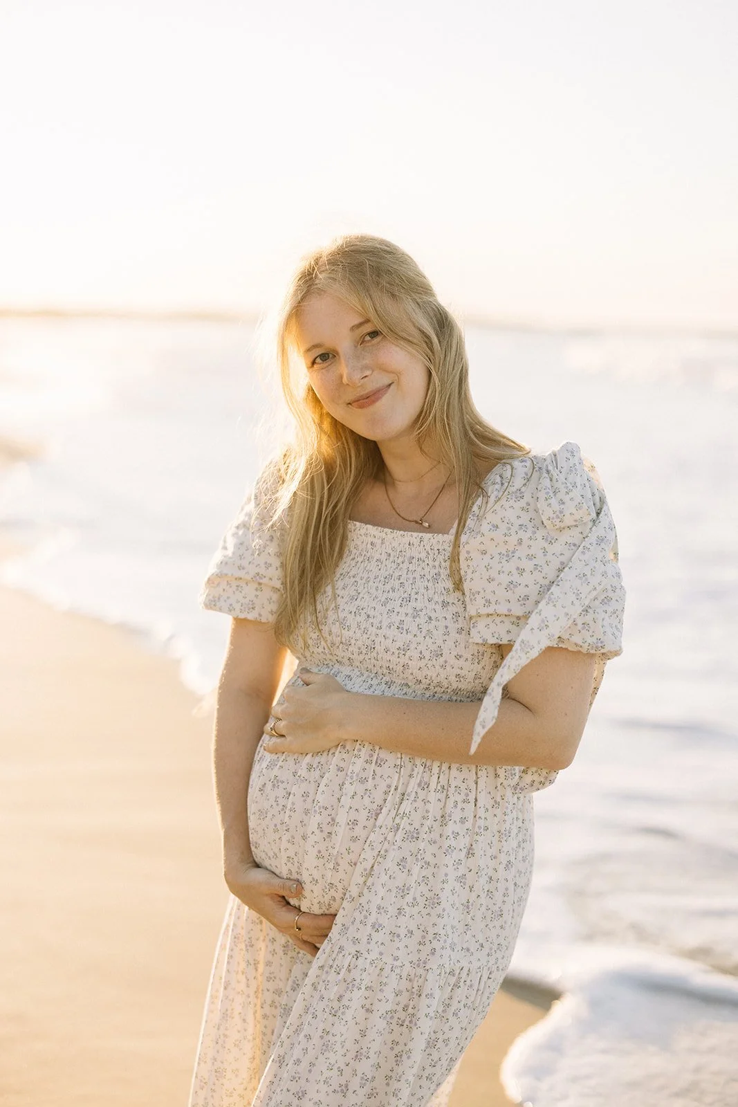A pregnant woman in a floral dress smiling and gently holding her belly on the beach during sunset. Taken by Wylde Folk Studio, Brisbane. Maternity photographer.