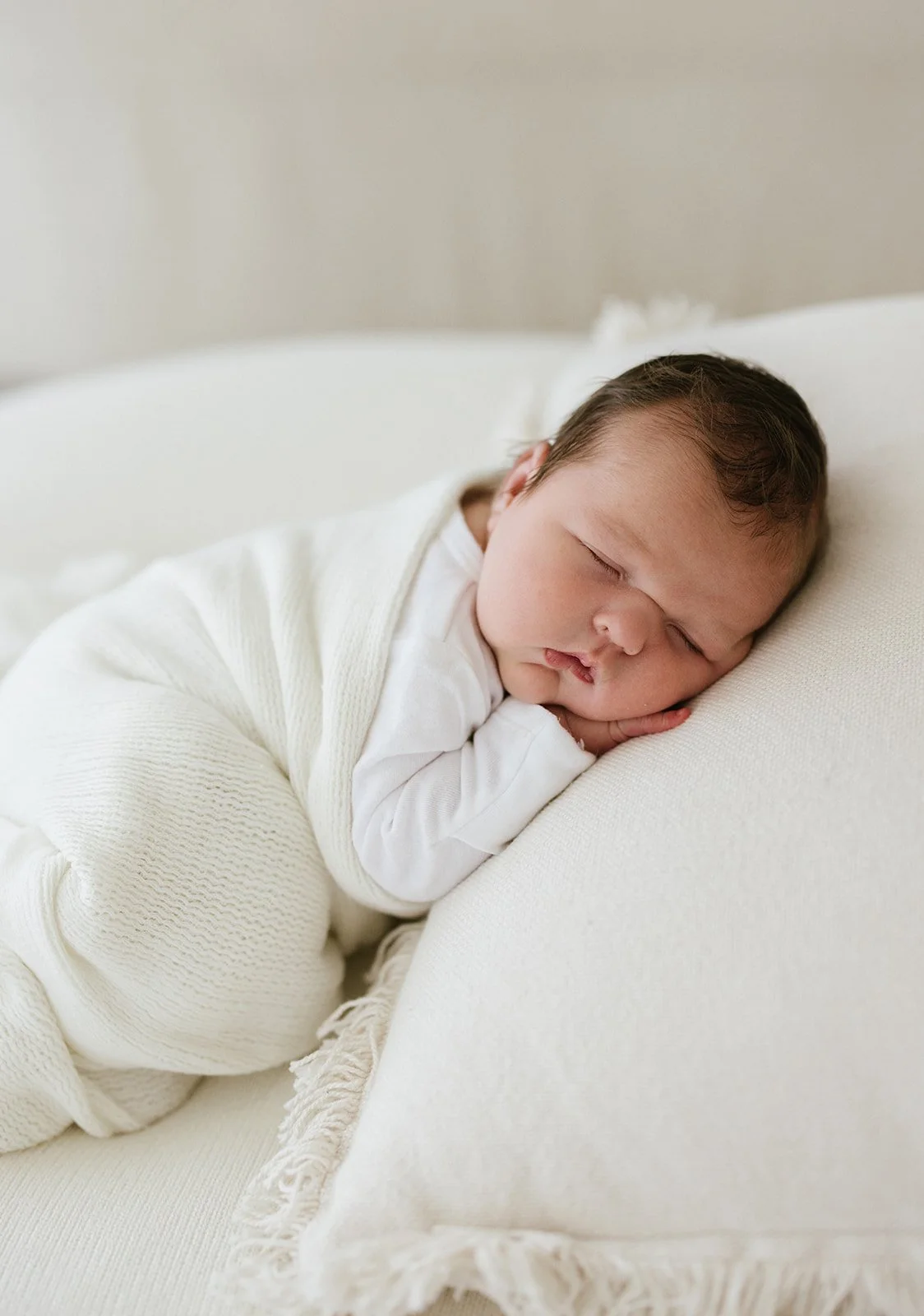 A sleeping baby with dark hair, resting on a white pillow, dressed in white clothing, lying on a cream-colored bed.