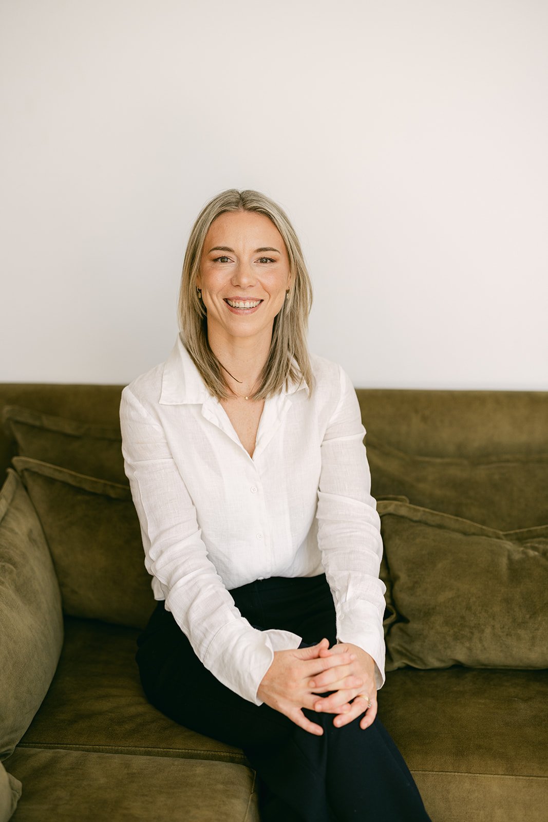 a blonde woman in professional attire is sitting on a dark green sofa posing for a professional head shot