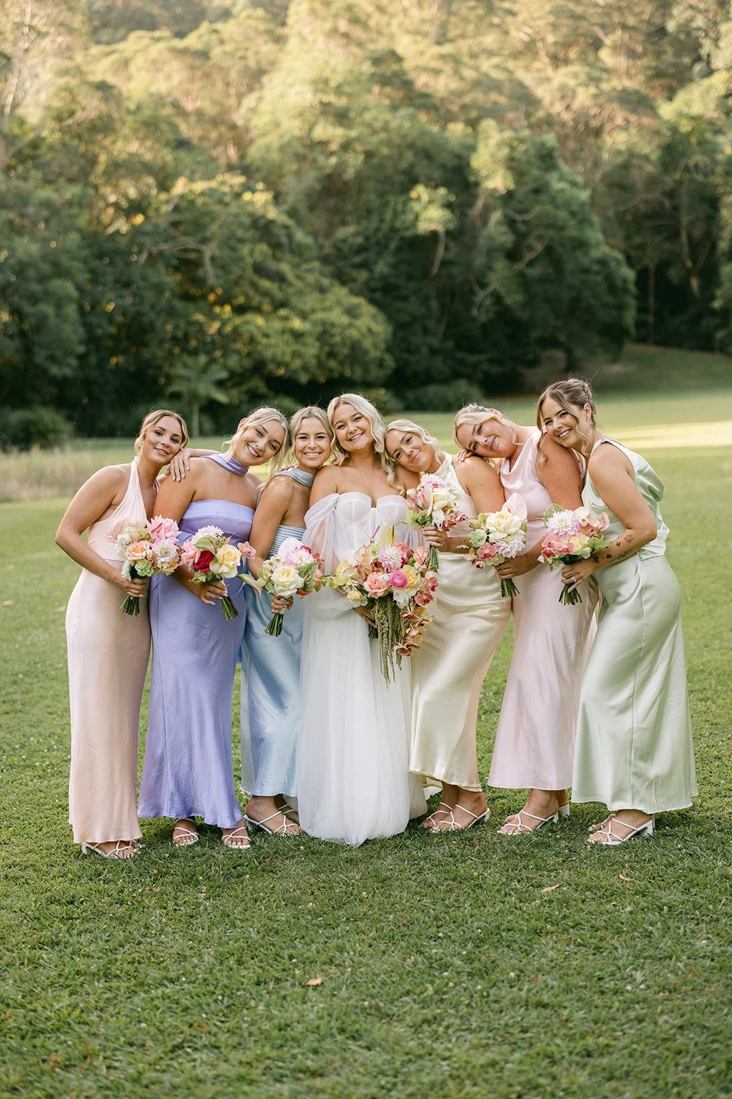 A bride and six bridesmaids stand together outdoors on a grassy field, smiling and holding bouquets of pink and white flowers. The bridesmaids wear pastel-colored dresses, and the bride is in a white dress, with a background of green trees.