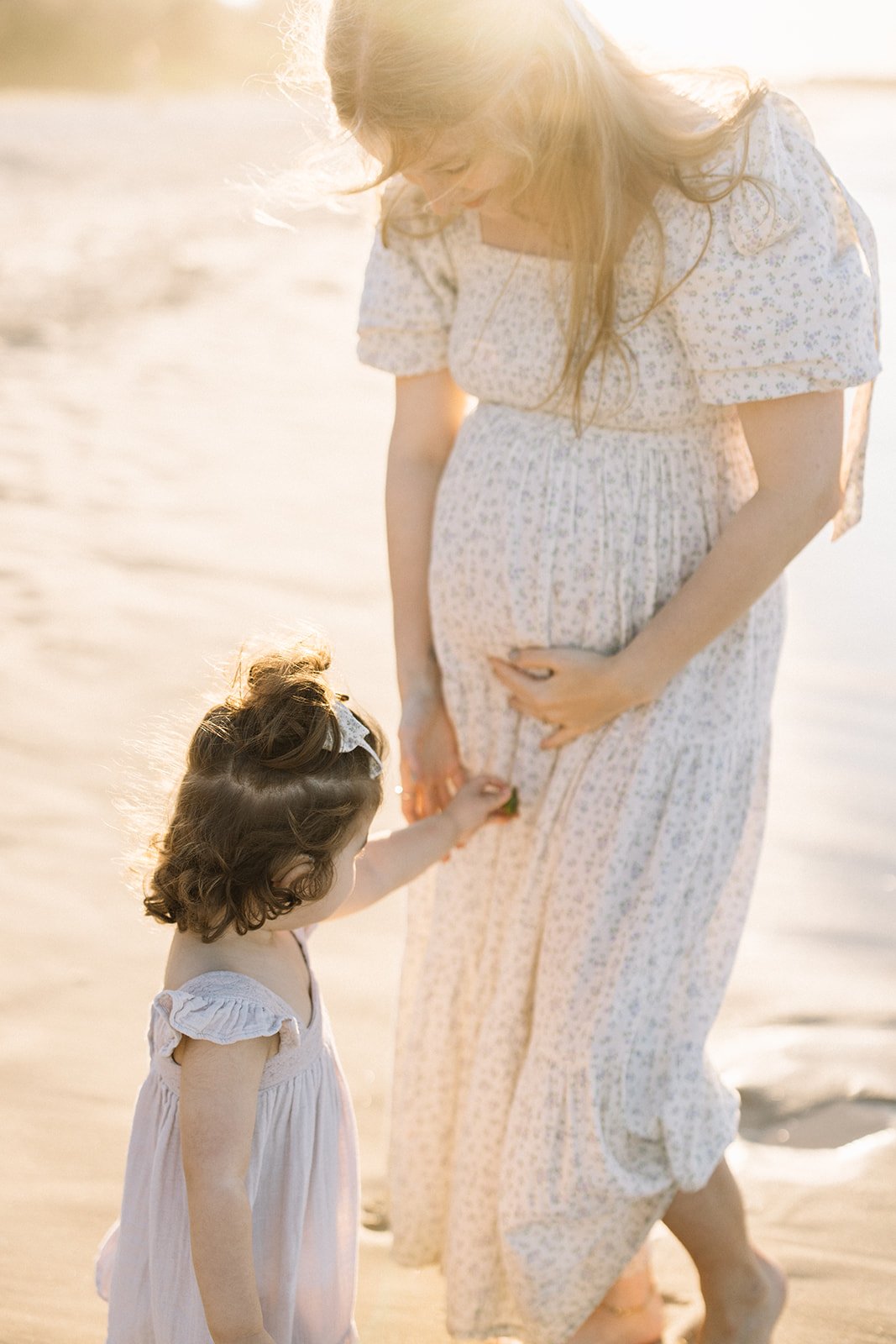 A pregnant woman in a white floral dress gently touches her belly while standing on the beach with a young girl, possibly her daughter, holding her hand. Taken by Wylde Folk Studio, Brisbane. Maternity photography.