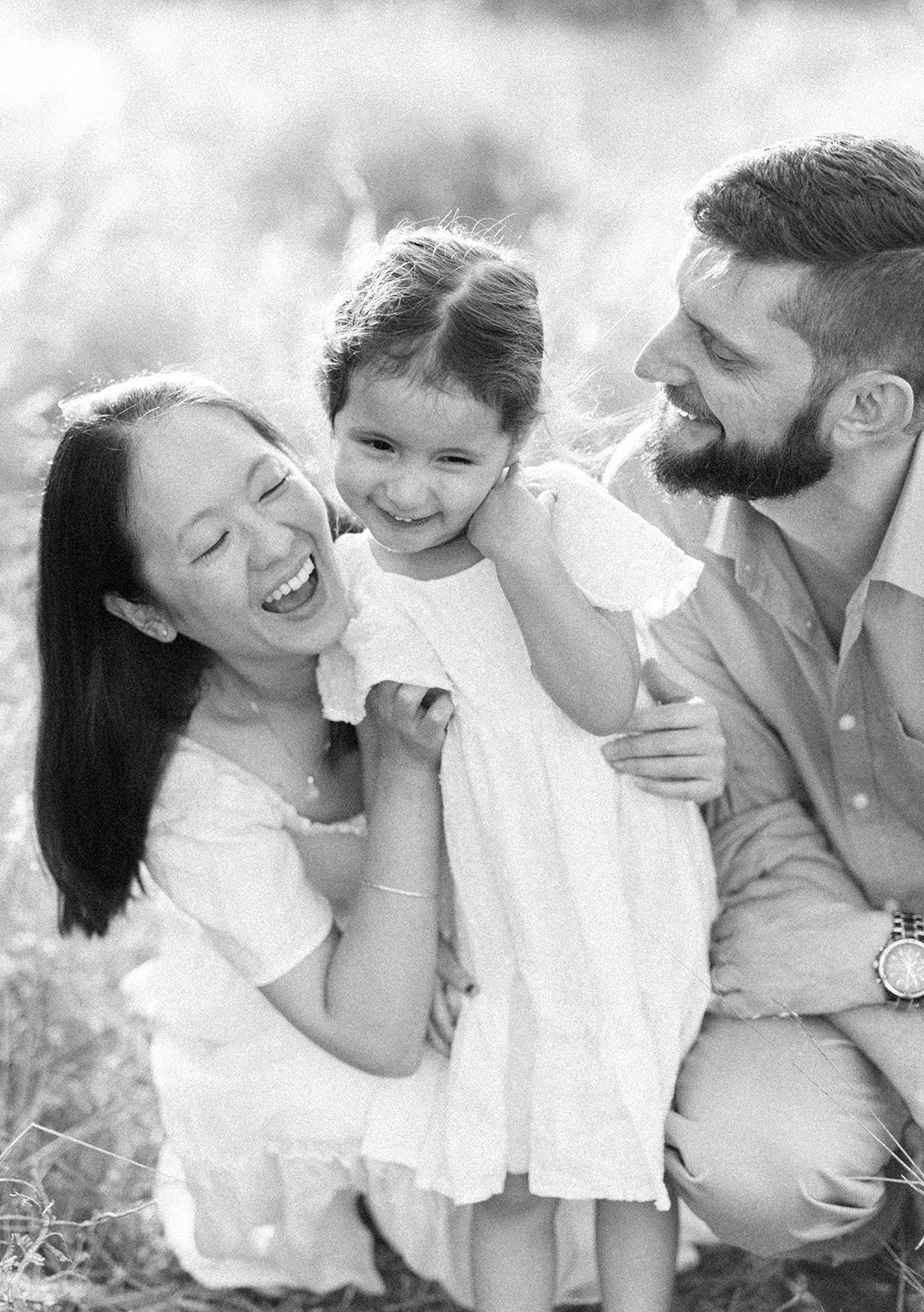 A happy family outdoors in a black and white photo, with a woman and a man smiling at a young girl they are holding. Taken by Wylde Folk Studio, Brisbane. Family photographer.