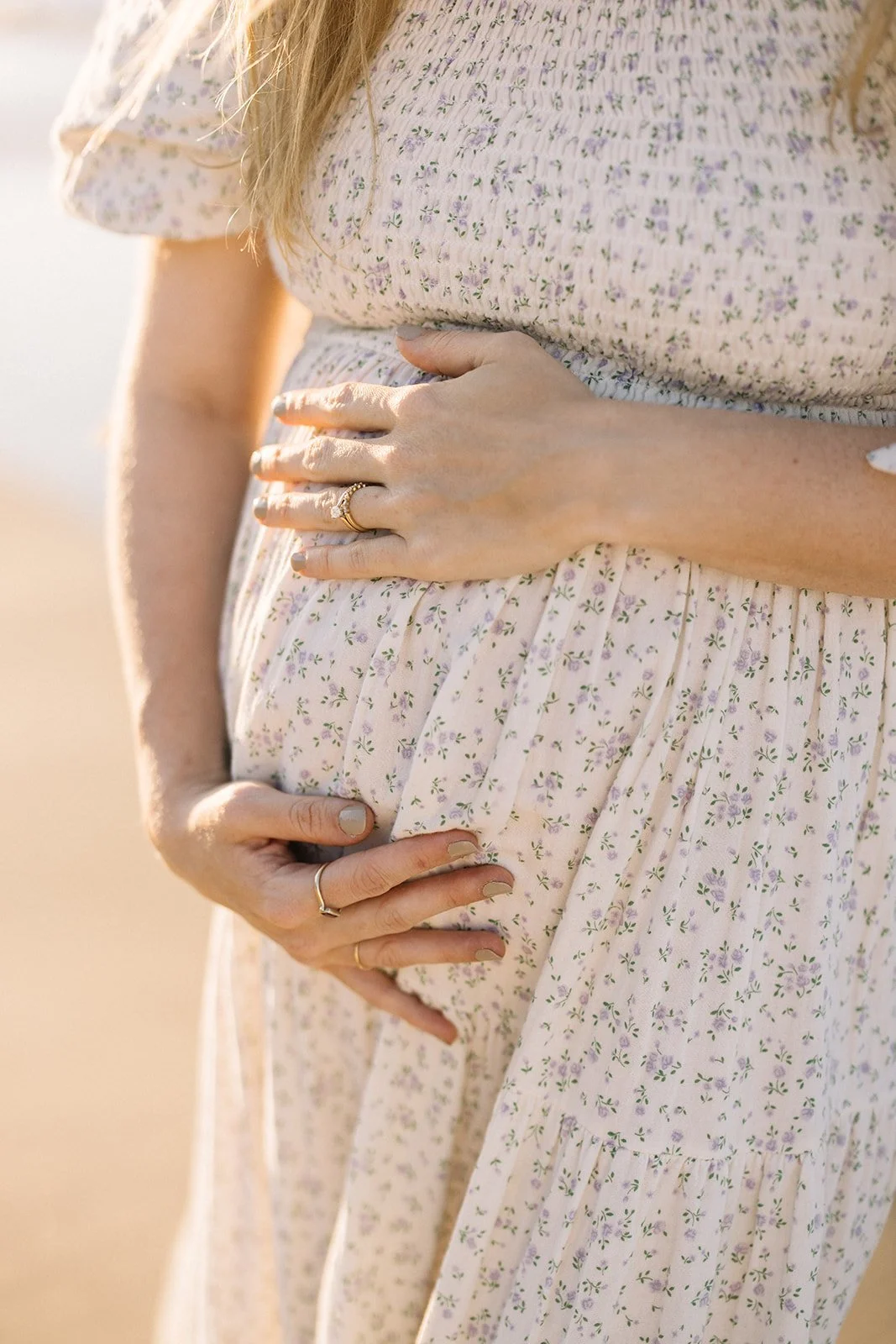 A pregnant woman in a light pink floral dress gently holds her belly with both hands, showcasing rings on her fingers. The scene has warm, natural lighting. Taken by Wylde Folk Studio, Brisbane. Maternity photography.