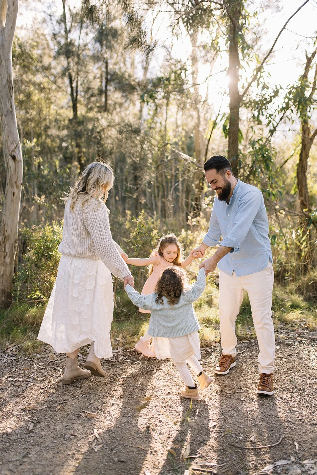 Family of four holding hands and dancing outdoors on a dirt path in a wooded area during golden hour. Taken by Wylde Folk Studio, Brisbane. Family photographer.