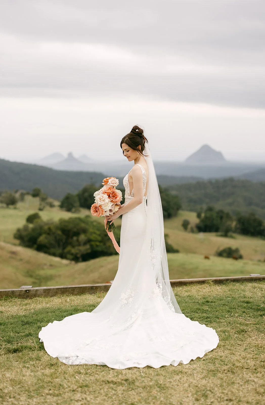 Bride in a wedding dress holding a bouquet of pink and cream roses outdoors on a cloudy day with rolling green hills and mountains in the background.