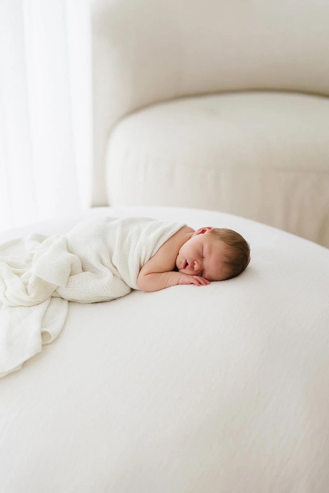 A newborn baby with brown hair lays on a white newborn photography beanbag on their tummy, the baby is sleeping peacefully with their little hand under their head. The photograph is by Brisbane newborn photography studio Wylde Folk Studio 