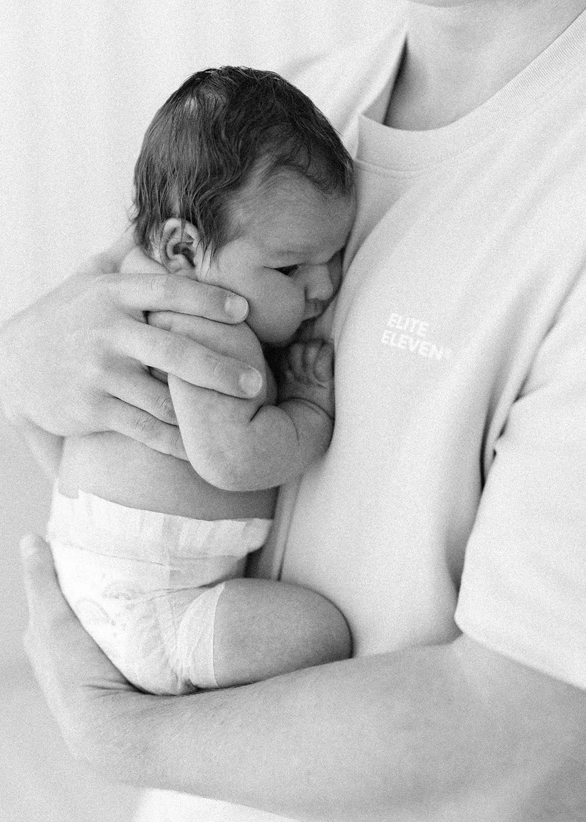 A newborn baby being held close to a person's chest, with the baby’s face resting on the person's breast, breastfeeding. The person is wearing a shirt with the words 'COTE ELEVEN' on it.