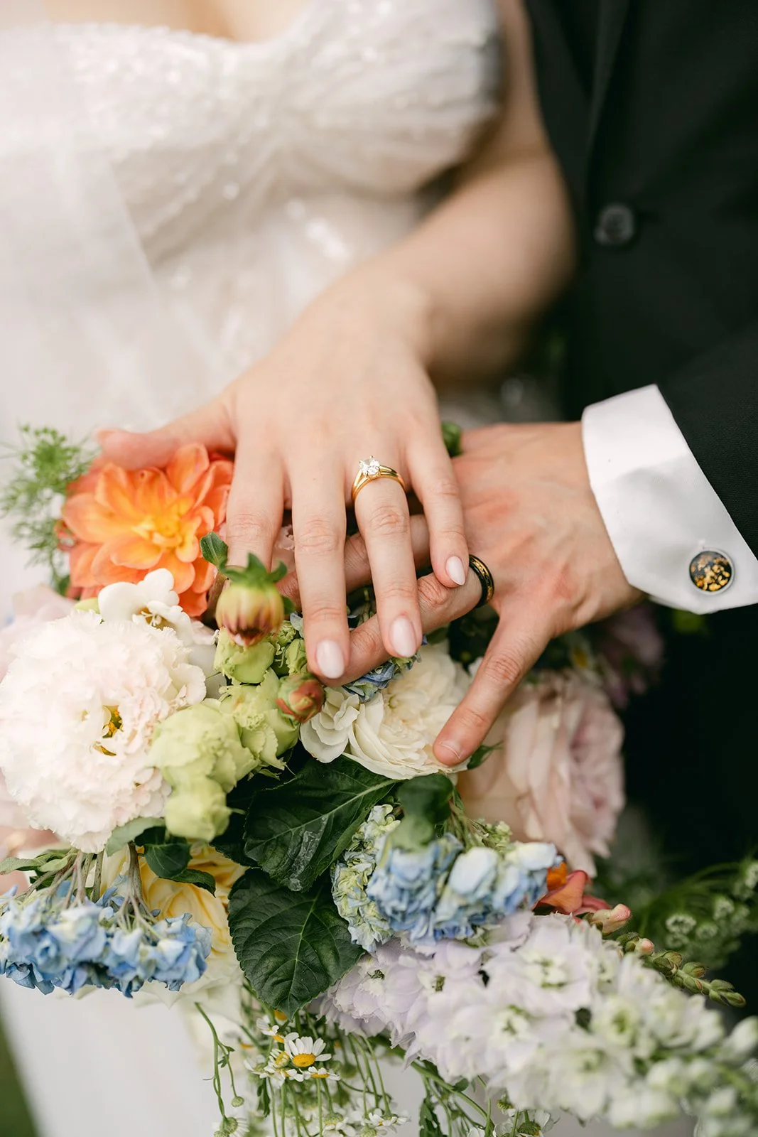 Close-up of a bride and groom's hands showing wedding rings, with a bouquet of colorful flowers including peonies, roses, and delphiniums.
