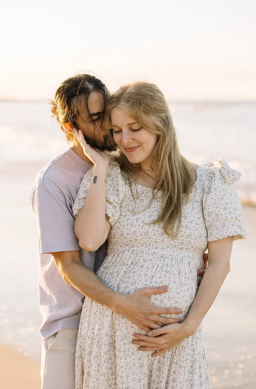 A pregnant woman and her partner embrace on a beach during sunset, with the man whispering into her ear and both smiling softly. Taken by Wylde Folk Studio, Brisbane. Maternity photography.
