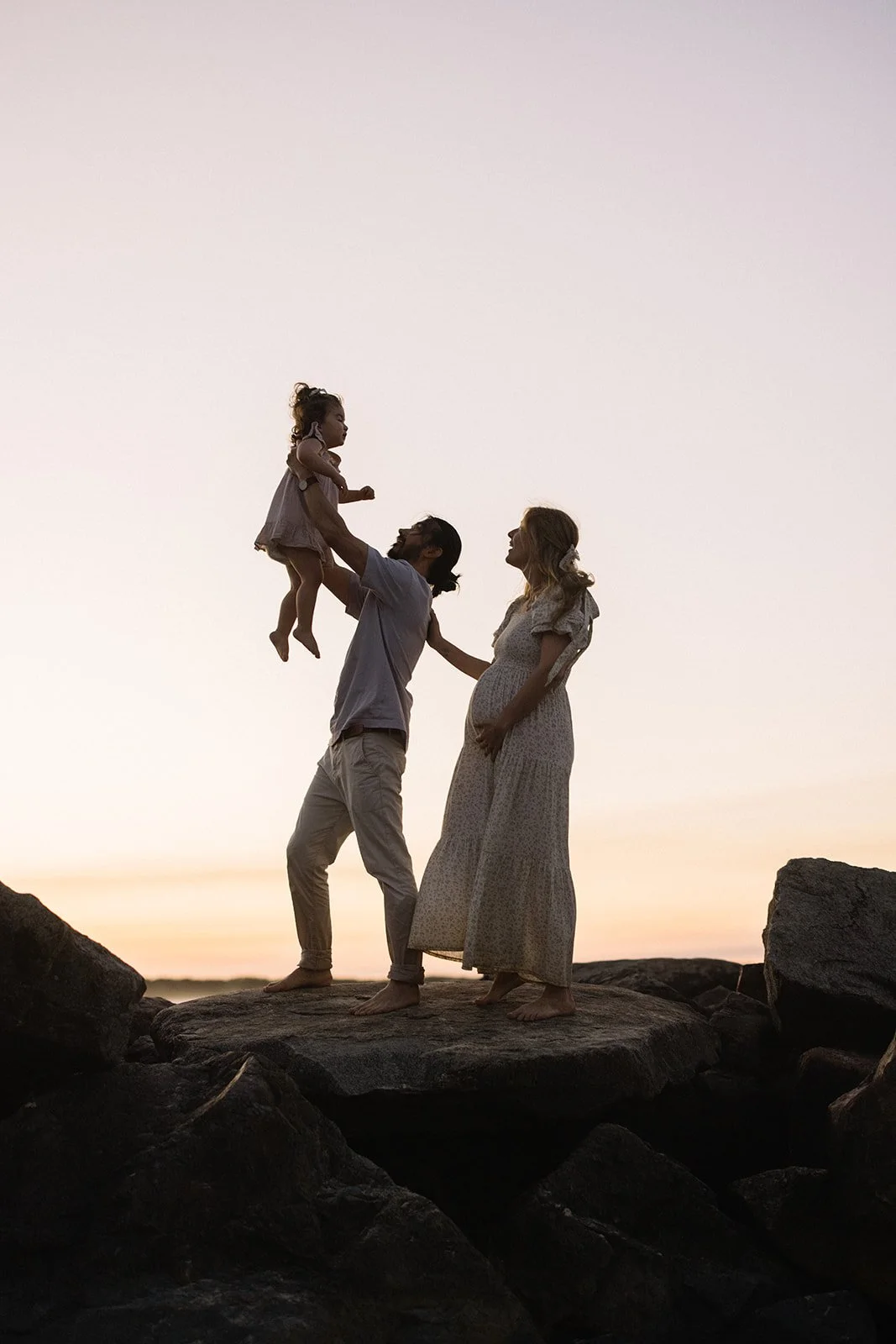 A family standing on rocks at sunset; the father lifts a young girl, and the mother touches her pregnant belly, all silhouetted against the sky. Taken by Wylde Folk Studio, Brisbane. Maternity photography.