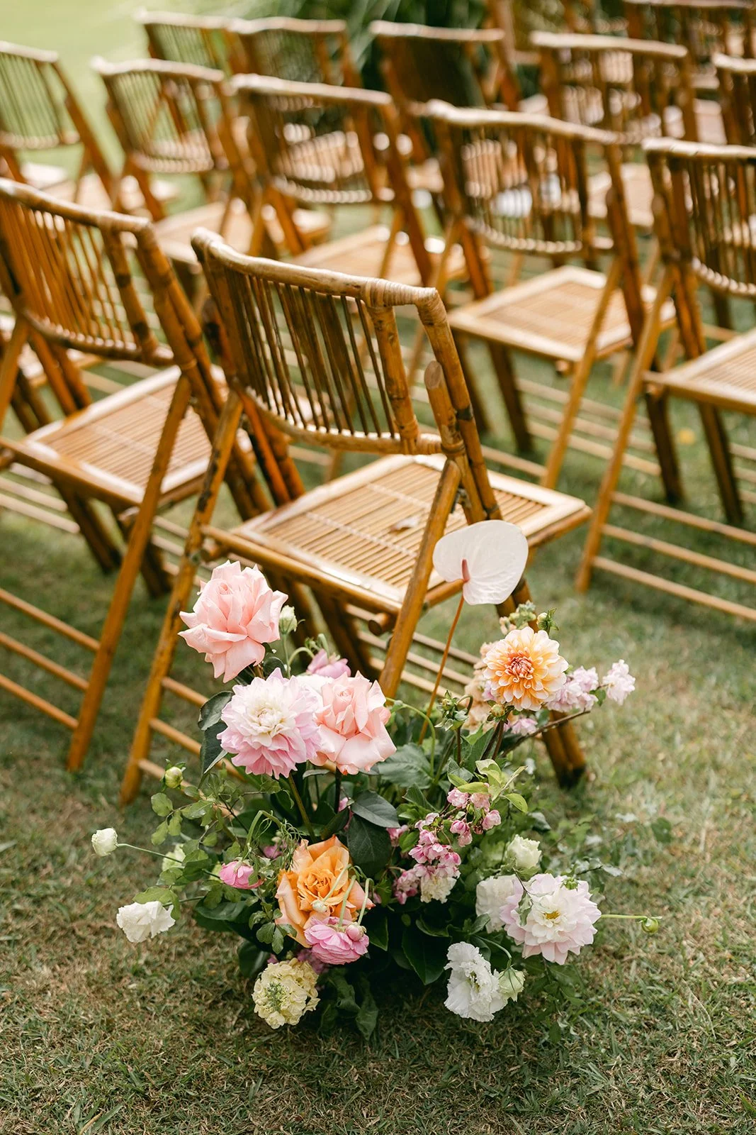 Empty wooden chairs arranged in rows for an outdoor event, with a large floral arrangement of pink, peach, and white flowers on the grass in front.