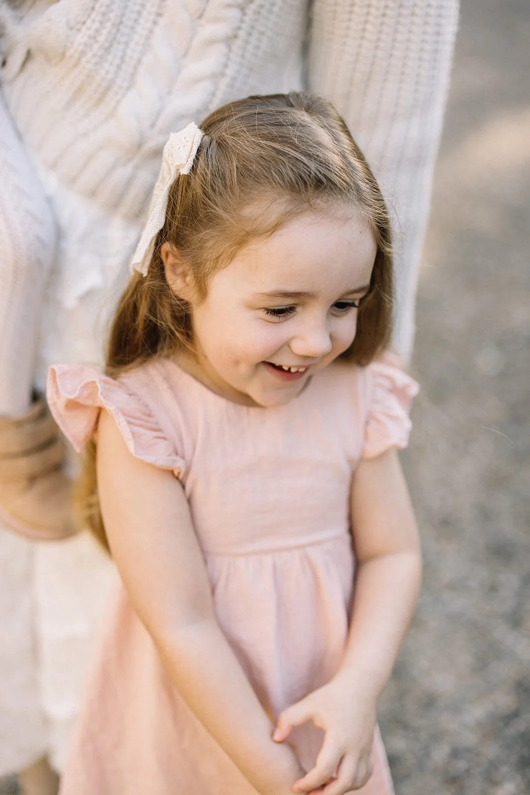 A young girl with light brown hair and a white hair bow, smiling and looking down, wearing a pink dress with ruffled sleeves, standing outdoors near a wooden structure. Taken by Wylde Folk Studio, Brisbane. Family photography.