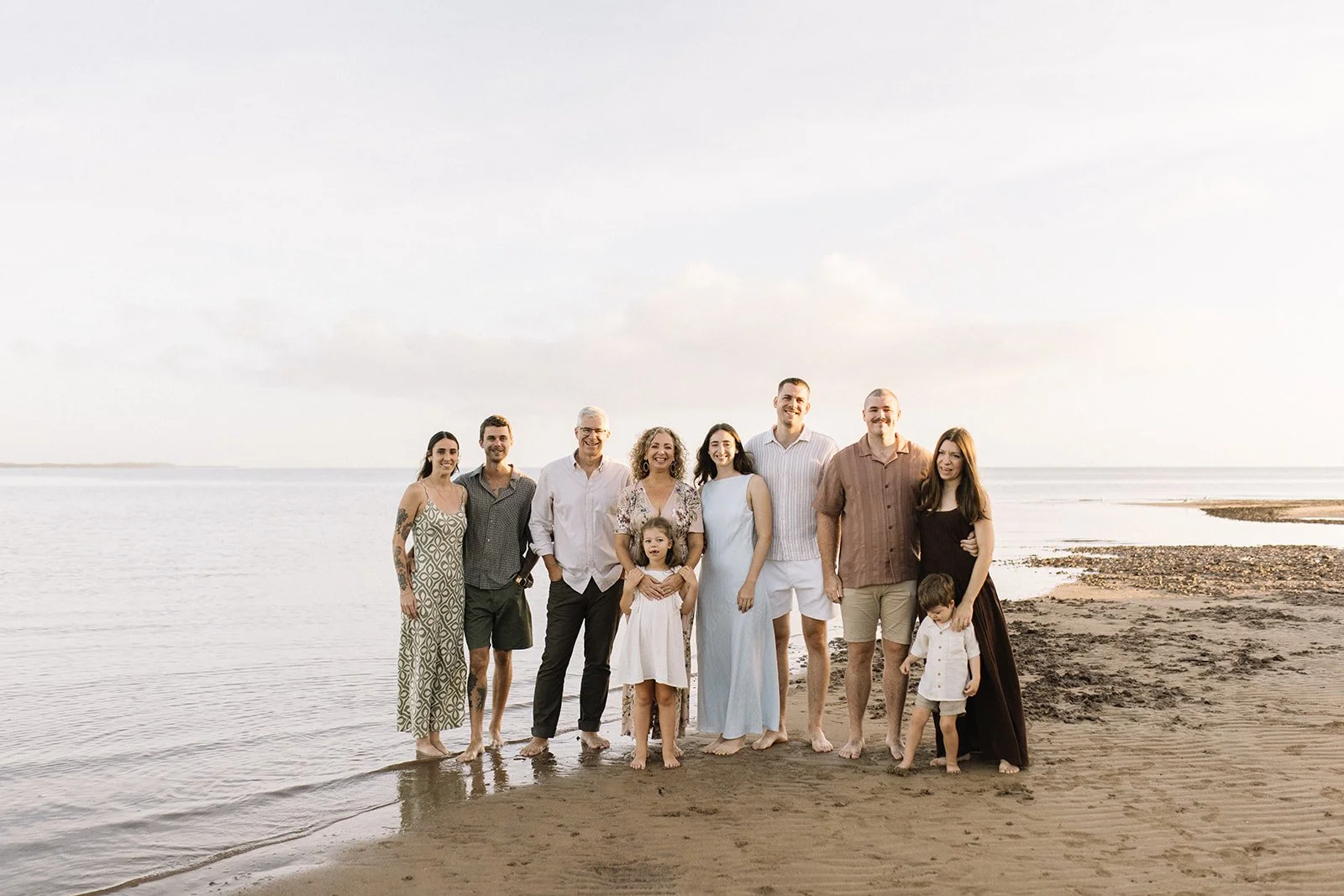 Group of adults and children standing barefoot on a beach by the water during sunset. Taken by Wylde Folk Studio, Brisbane. Family photographer.