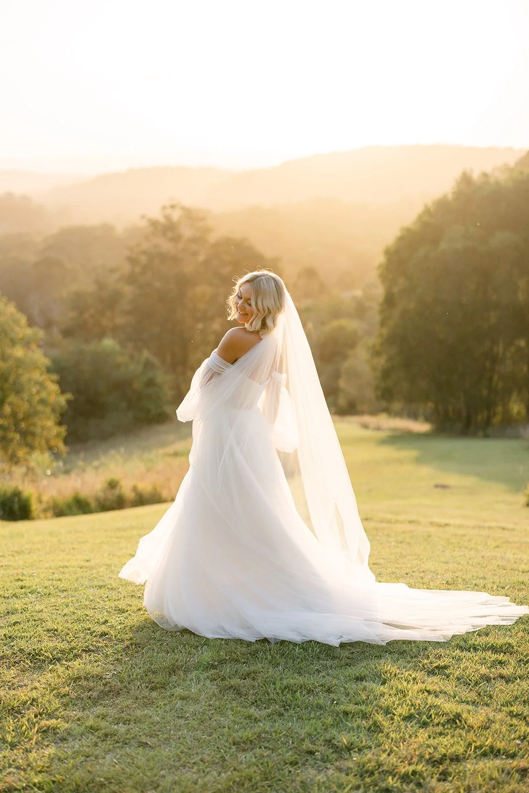 A woman in a wedding dress standing in a grassy field during sunset, smiling with her eyes closed.