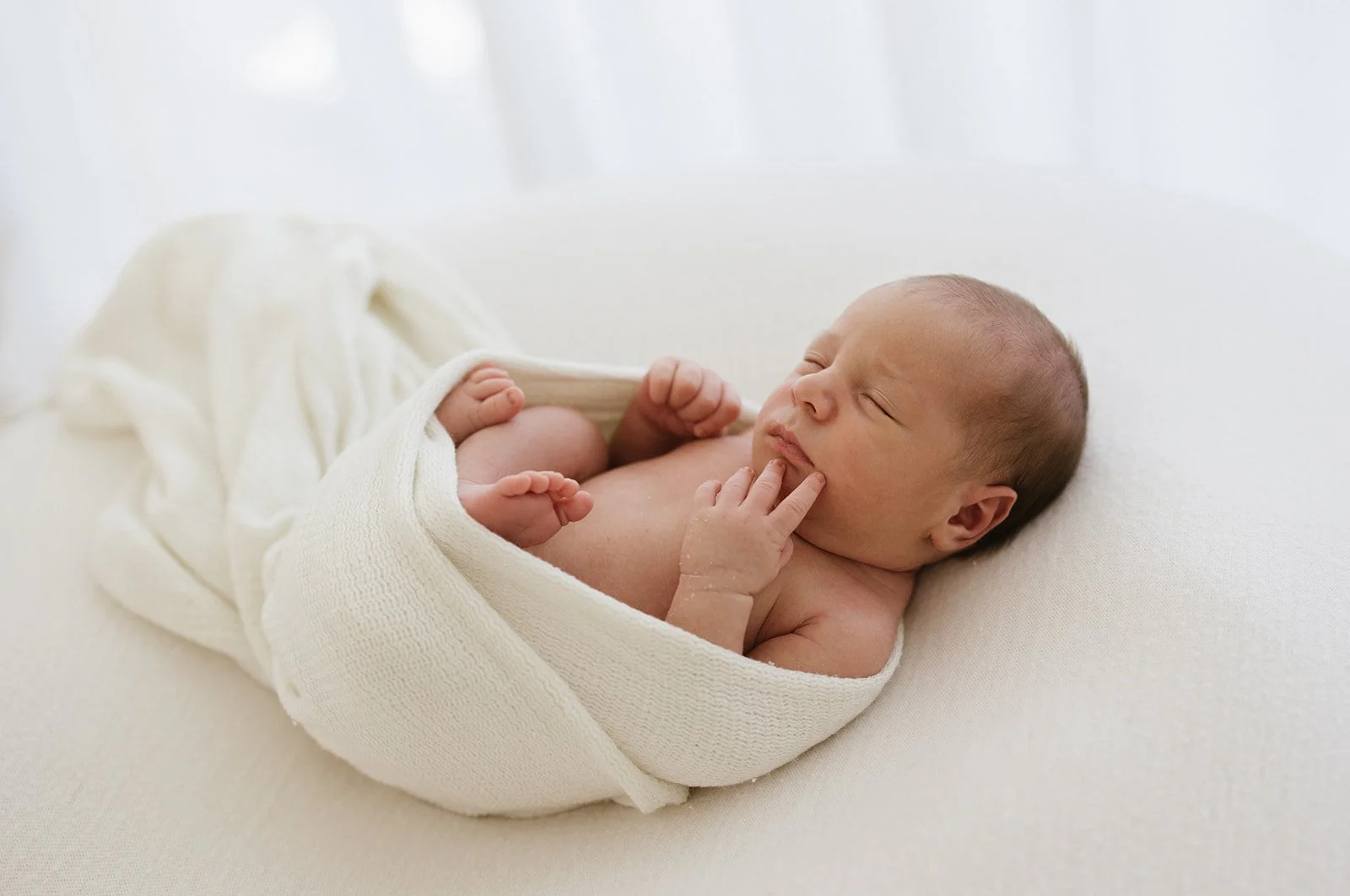 A sleeping newborn baby is swaddled in a white blanket, lying on a white cushion