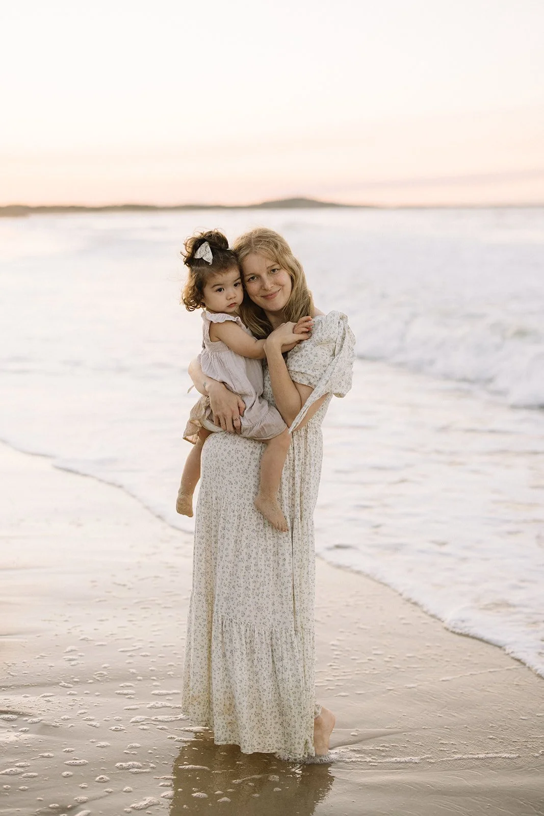 A woman holding a young girl while standing on the beach at sunset, with the ocean and sky in the background. Taken by Wylde Folk Studio, Brisbane. Maternity photography.