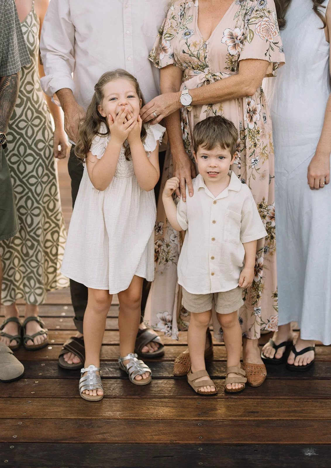 Two young children, a girl and a boy, standing on a wooden dock, dressed in light-colored summer clothing, surrounded by adults at what appears to be a family gathering or celebration.