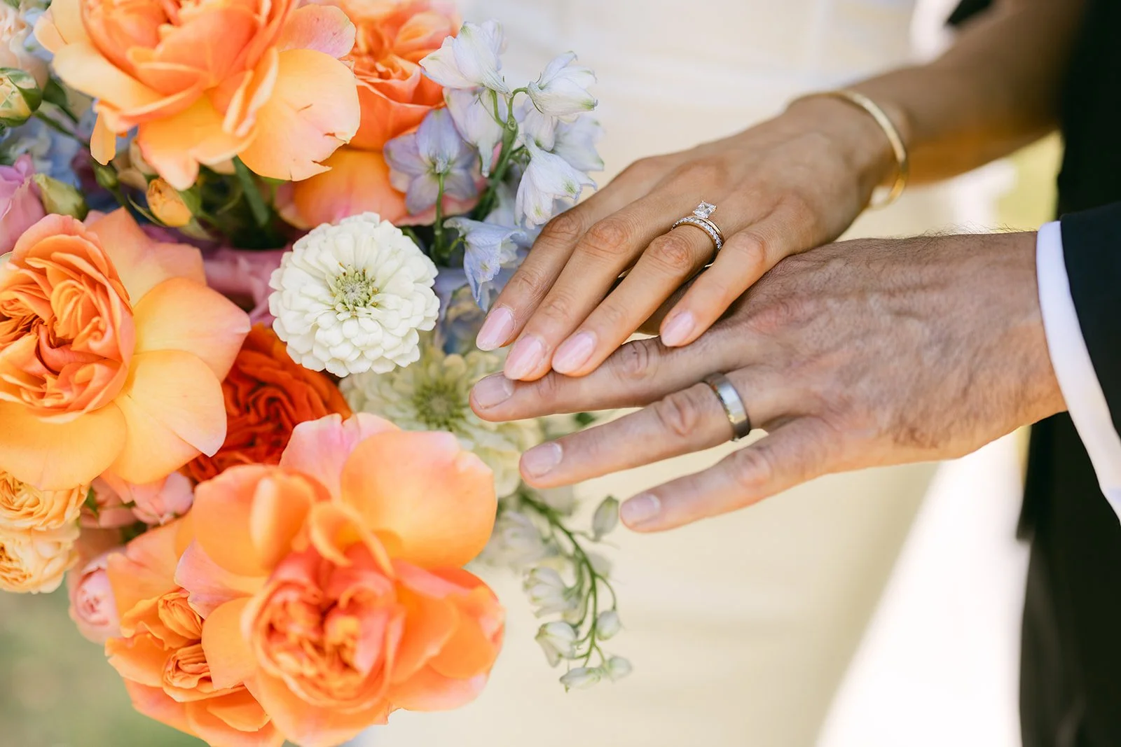 Close-up of a married couple's hands showing wedding rings, resting on a bouquet of orange, peach, white, and purple flowers, with a blurred background.