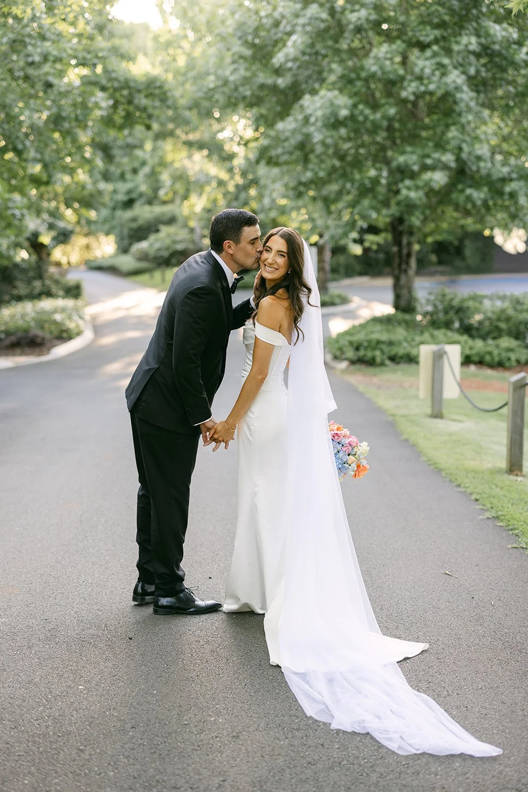 A newlywed couple, dressed in wedding attire, standing on a paved pathway in a park-like setting with green trees and sunlight filtering through, holding hands and sharing a kiss, with the bride smiling happily and holding a colorful bouquet of flowe