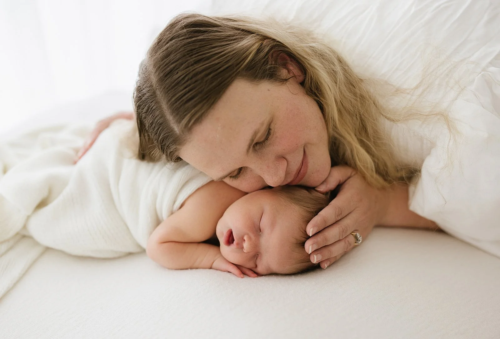 Newborn baby sleeps on white beanbag on their tummy. Mother of baby gently rests her head on baby's head while cuddling them.