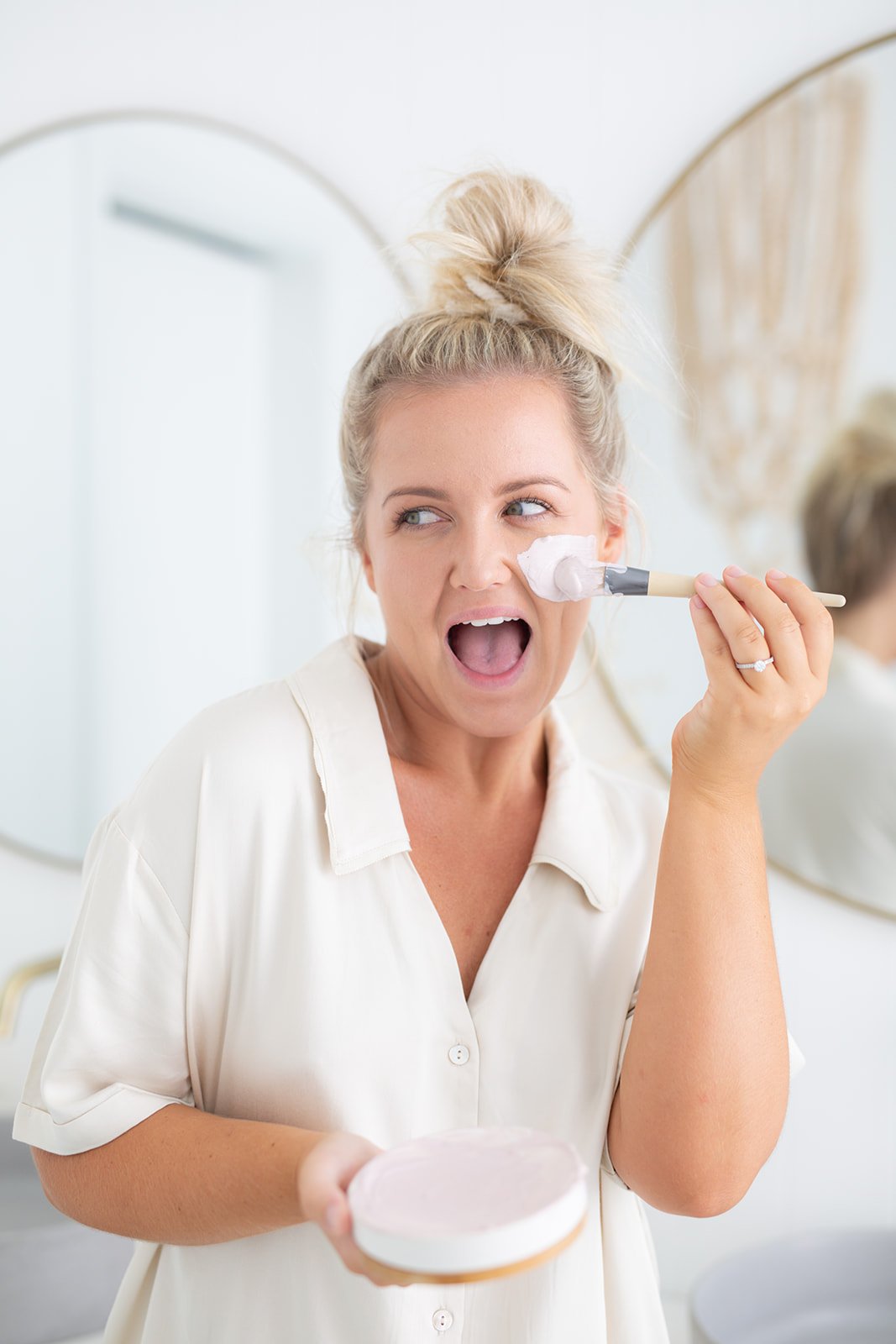 A blonde woman with her hair in a bun is applying a face mask to her cheek while smiling with her mouth open