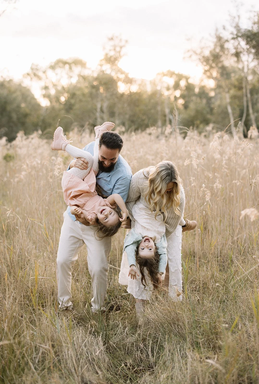 A family of four playing and laughing in a grassy field during sunset, dad holding daughter upside down, while mom and another daughter are smiling and bending down.