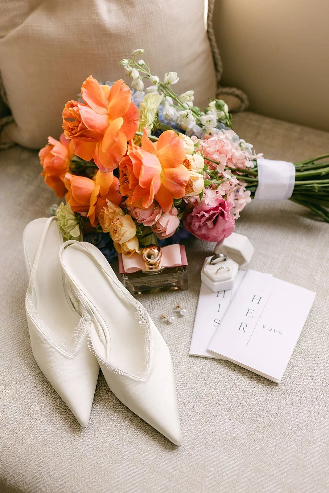 A pair of white pointed-toe high heels, a bouquet of pink, orange, and white flowers, a small perfume bottle with a pink bow, a jewelry box with rings, pearl earrings, and wedding vows on paper reading "HIS" and "HER" on a beige fabric surface.