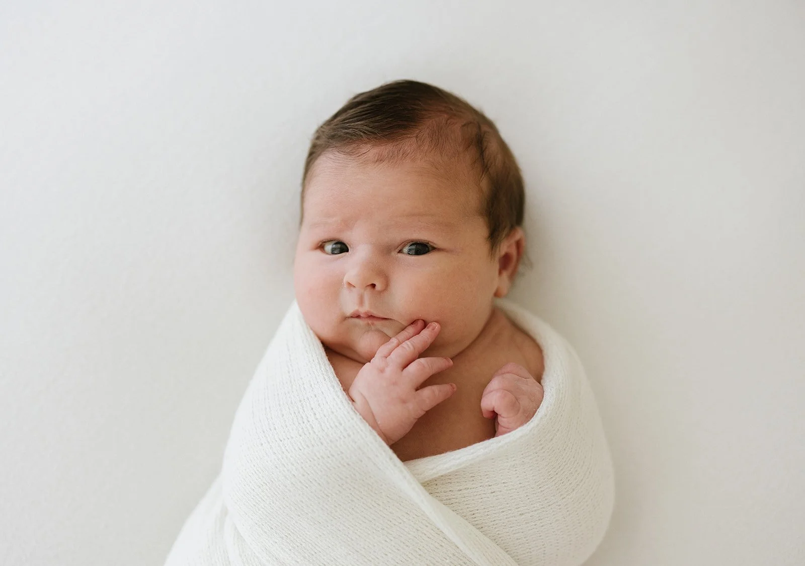 Close-up of a newborn baby wrapped in a white blanket with a thoughtful expression, against a plain white background.